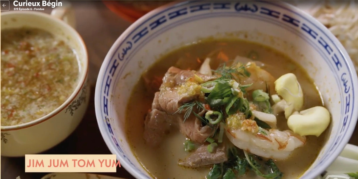 A close-up of a bowl of soup with meat, garlic, green onions, and herbs, alongside a smaller bowl of a lighter-colored soup. The bowls have blue and white patterns, and the scene has a warm, restaurant-like feel.